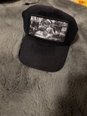 Svart truckerkeps från Maggiore - Svart truckerkeps från Maggiore med mesh på sidorna och justerbart spänne bak. Framtill finns en svartvit patch med motiv av en racerförare. Kepsen har en böjd skärm och metallogo på sidan. Perfekt för dig som gillar motorsport och streetwear.