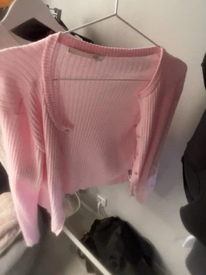 Rosa ribbad kofta med knappar - Säljer en ljusrosa kofta med ribbad struktur och rund hals. Koftan har långa ärmar och stängs med små knappar framtill. Den är croppad i modellen och gjord i ett mjukt stickat material som känns skönt mot huden. Perfekt för att addera lite färg till din outfit.