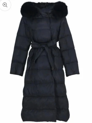 Svart lång pufferjacka STORLEK S - Säljer en svart lång pufferjacka med stor huva och pälsdetalj. Jackan har bälte i midjan för snygg passform och är quiltad för extra värme. Perfekt för kalla dagar och ger en cool och trendig look.