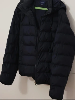 Svart pufferjacka från GANT - Säljer en mörkblå pufferjacka / vinterjacka från GANT med huva och dragkedja framtill. Den har en klassisk look och passar dig som gillar stilrena och funktionella jackor.