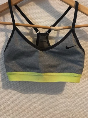 Grå och gul sport-bh från Nike - Sport-bh från Nike i grått med svarta detaljer och en gul elastisk kant nedtill. Snygg design med tunna axelband och meshparti i ryggen. Perfekt för träning och aktiva dagar. Nike-logga på framsidan.