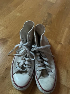 Vita Converse (höga) - Klassiska höga vita Converse  med röd rand runt sulan och ikonisk logga på sidan. Skorna har snörning, rund tå och vit gummisula. Perfekta för en avslappnad och tidlös stil.