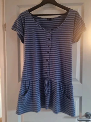 Blå och grå randig kortklänning - Säljer en blå och grå randig kortklänning från Only med knappar framtill, fickor och knyt i midjan. Klänningen har korta ärmar och är gjord i mjuk bomull, perfekt för en chill och avslappnad stil.