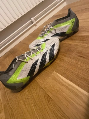 Adidas fotbollsskor i vitt och neon - Säljer ett par Adidas fotbollsskor med snygg design i vitt, grått och neonlime. Skorna har svarta ränder på sidorna, snörning och mönstrad ovandel. Yttersulan har dobbar för bra grepp på planen. Perfekt för dig som vill sticka ut på matchen!