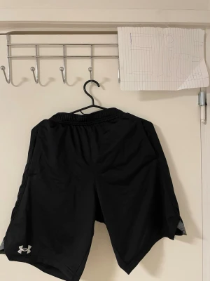 Svarta träningsshorts  Under Armour - Säljer ett par svarta träningsshorts från Under Armour med elastisk midja i grått och logga i vitt på vänster ben. Shortsen har loose passform och är gjorda i ett lätt syntetmaterial som andas bra. Perfekta för gymmet eller löprundan.Liknande modell som originala.