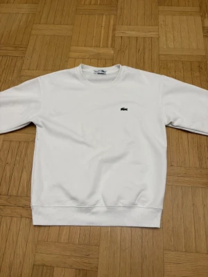 Vit sweatshirt från Lacoste - Säljer en stilren vit sweatshirt från Lacoste med klassisk rund hals och det ikoniska gröna krokodilmärket broderat på bröstet. Tröjan har långa ärmar och ribbade muddar vid hals, ärmslut och nederkant. Perfekt för en clean och sportig look.