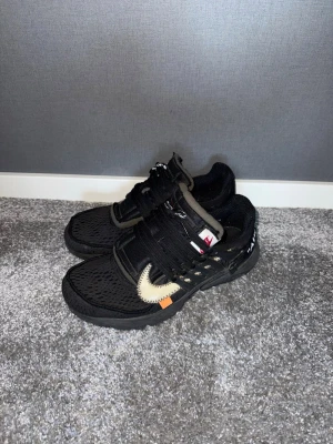 Nike Presto Off-White  - Nike Presto Off-White sneakers i svart mesh med ikonisk vit Swoosh och orange detalj. Skorna har snörning, platt sula och coola Off-White detaljer som synliga sömmar och text på sidan. Perfekt för dig som gillar streetwear och unika collabs. Väl använda