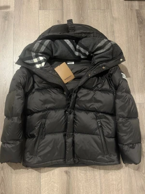 Burberry jacka / väst - Svart pufferjacka från Burberry med avtagbara ärmar, så du kan bära den som väst. Jackan har dragkedja, knappar och två fickor med zip. Insidan av huvan har ett grått och vitt rutigt mönster. Stor Burberry-patch på ärmen.