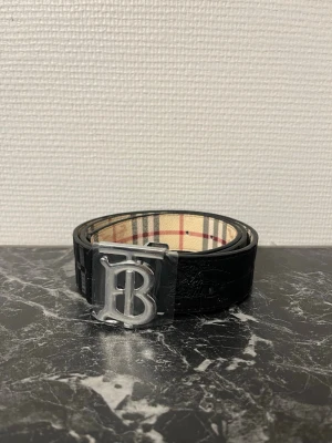 Rutig och svart bälte - Säljer ett bälte med vändbar design – ena sidan är klassiskt rutig i beige, svart och röd, andra sidan är helsvart med präglat mönster. Bältet har en stor silvrig metallspänne med 'B'-logga. Perfekt accessoar för att lyfta din outfit.