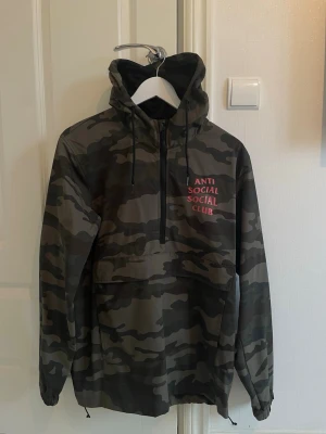 Camouflage vindjacka Anti Social Social Club - Säljer en snygg camouflage vindjacka från Anti Social Social Club med stor rosa logga på ryggen och mindre tryck på bröstet. Jackan har huva, half zip och stor ficka framtill. Perfekt för dig som gillar streetwear och vill sticka ut.