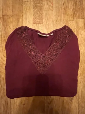 Vinröd topp med spets från Gina Tricot - Snygg vinröd långärmad topp från Gina Tricot i storlek 146/152. Toppen har en v-ringad hals med dekorativ spetsdetalj framtill. Materialet känns mjukt och bekvämt, perfekt för dig som gillar stilrena plagg med en twist.