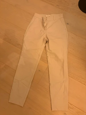 Beige kostymbyxor från H&M, strl 36 - Snygga beige kostymbyxor från H&M i storlek 36. Byxorna har hög midja, raka ben och bälteshällor. De är gjorda i ett mjukt bomullsmaterial med diskreta bakfickor och dragkedja fram. Perfekt för en clean och stilren look. Mindre märke på baksidan, se sista bilden.
