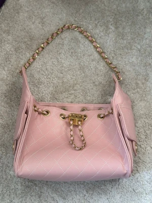 Rosa quiltad - Ch@nel 25 - Säljer en rosa axelväska från Ch@nel 25 i skinn med quiltat mönster och guldfärgade kedjedetaljer. Väskan har sidofickor med lock och ikoniska CC-loggan i guld både fram och på fickorna. Insidan har dragkedja och snygga detaljer. Perfekt för dig som vill sticka ut med lyxig stil.