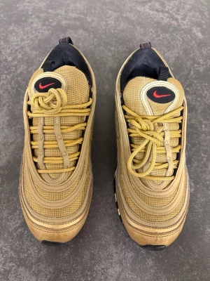 Nike Air Max 97 Gold sneakers - Nike Air Max 97 sneakers i metallic guld med röda swoosh-detaljer. Skorna har en vågig design, synlig Air-sula och snörning framtill. Ovandelen är i syntet och mesh, med svarta och vita inslag på sula och foder. Perfekt för dig som gillar att sticka ut med din stil.