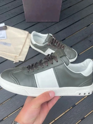 Valentino fly crews grön/vit - Snygga Valentino Garavani sneakers i grått och vitt skinn med bruna snören och coola nitar på sulan. Sulan har ett unikt camouflage-mönster undertill. Klassisk rund tå och platt sula, perfekt för dig som gillar stilrena och exklusiva sneakers. Skorna är näst intill oandvända och i toppskick. Allt og medföljer.dvs box, påsar, kvitto och nya snören.