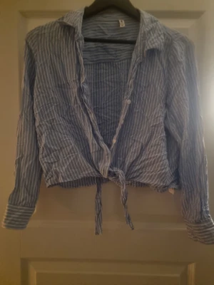 Blåvit randig skjorta med knyt - Blå skjorta med vita ränder och långa ärmar. Skjortan har knappar framtill och en snygg knytning nedtill för en avslappnad look. Tillverkad i ett lätt och mjukt material, perfekt för varmare dagar. Klassisk krage och manschetter med knapp.