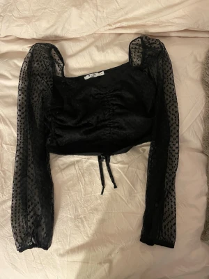 Svart prickig blus från NA-KD - Säljer en svart blus från NA-KD i storlek M. Toppen har transparenta långärmade meshärmar med prickigt mönster och en croppad passform. Framtill finns en snygg snörning som ger extra detalj. Perfekt för dig som gillar trendiga och unika plagg. endast använda en gång