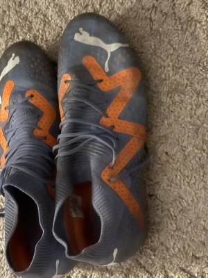 Blå-orange fotbollsskor från Puma - Säljer ett par fotbollsskor från Puma i blått med orange detaljer och vita Puma-loggor. Skorna har snörning och plös i stickad syntet, samt orange dobbar på sulan för bra grepp på planen. Perfekt för dig som vill sticka ut med färg och stil på fotbollsplanen.