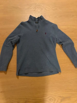 Blå half zip tröja från Polo Ralph Lauren - Säljer en stilren blå långärmad tröja från Polo Ralph Lauren i storlek S. Tröjan har en half zip vid halsen och klassisk röd broderad logga på bröstet. Materialet är mjukt och känns som bomull, perfekt för dig som gillar enkel och snygg design.