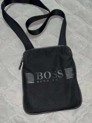 Svart axelväska från Hugo Boss - Snygg svart axelväska från Hugo Boss med justerbar rem och dragkedja upptill. Väskan har ett stilrent tryck med BOSS-loggan på framsidan och är tillverkad i slitstark polyester. Perfekt storlek för mobil, plånbok och andra småsaker.
