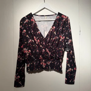 Svart omlottopp med blommigt mönster - Säljer en svart omlottopp från Nelly med blommigt mönster. Toppen har lång ärm, v-ringad omlottfront och smockad midja som ger en snygg croppad look. 