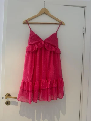 Rosa kort klänning med volanger - Säljer en söt rosa kort klänning med tunna axelband och volangdetaljer. Klänningen har prickigt mönster och är tillverkad i ett lätt, luftigt material som ger en skön känsla. Perfekt för varma dagar!!