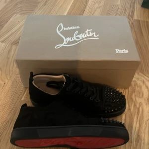 Svarta Christian Louboutins med nitar  - Säljer ett par svarta sneakers från Christian Louboutin med klassisk röd sula och svarta nitar på tån. Skorna har snörning och är tillverkade i mocka med läderdetaljer på insidan. Perfekta för dig som vill sticka ut med en edgy stil. Storleken 40 och dom är större i storleken som passar 40,5