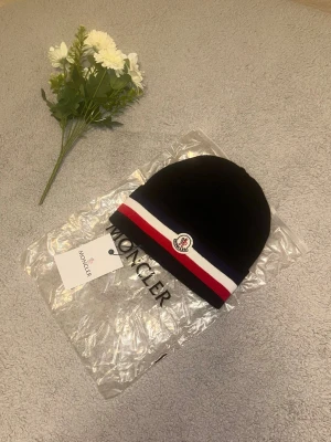 Svart Moncler mössa med ränder - Säljer en stilren beanie som endast har använts 1–2 gånger. Den är i utmärkt skick! Jag är aktiv 24/7 och kan frakta samma dag eller nästa dag. Om du har några frågor, tveka inte att höra av dig! Köp nu för att göra den till din egen!🚛✅📦