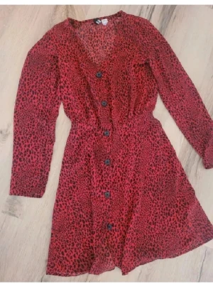 Röd leopardmönstrad kortklänning från H&M Divided - Snygg röd kortklänning med svart leopardmönster. Klänningen har v-ringning, långa ärmar och knappar framtill. Den är tillverkad i ett lätt och luftigt material som ger en skön känsla. Perfekt för dig som vill sticka ut med en cool look.