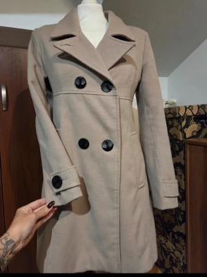 Beige kappa med svarta knappar storlek 38 - Snygg beige kappa med dubbelknäppning och stora svarta knappar. Kappan har klassisk krage, långa ärmar med knappdetalj vid mudden och två fickor framtill. Insidan är fodrad med svart tyg. Kappan är i en längre modell och går till halva låret på mig som är 156cm lång. Där är även lite vaddering i axlarna. Perfekt för dig som gillar stilrena och tidlösa plagg. Kappan är i otroligt fint skick ock knappt använd. Priset går att diskutera.