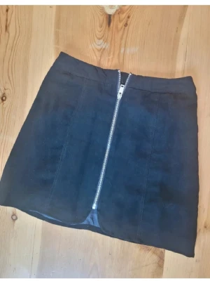 Svart kortkjol med dragkedja - Säljer en svart kortkjol med en snygg silverfärgad dragkedja framtill. Kjolen har en enkel och stilren design som gör den lätt att matcha med olika toppar. Perfekt för dig som gillar en modern och clean look.