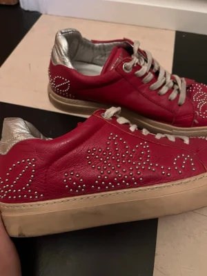 Röda Zadig & Voltaire sneakers med nitar - Snygga röda sneakers från Zadig & Voltaire med silverfärgade detaljer och coola mönster av små nitar på sidorna och tån. Skorna har snörning och en beige platt sula. Insidan är fodrad med metallic-silver och märket syns tydligt på plösen.