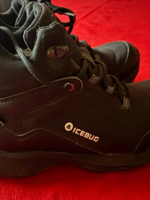 Svarta Icebug kängor med dubbar - Säljer ett par svarta Icebug kängor med grov sula och metall-dubbar för extra grepp. Skorna har snörning och är tillverkade i slitstark syntet och skinn. Perfekta för vinter och hala underlag, med coola gula detaljer på undersidan.