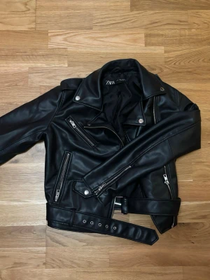 Svart skinnjacka från Zara, S - Snygg svart skinnjacka från Zara i bikerstil med flera dragkedjor, silvriga knappar och bälte i midjan. Jackan har klassisk krage och coola detaljer som ger en edgy vibe. Perfekt för dig som vill ha en tidlös och trendig look.