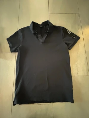 Svart piké från Peak Performance, 13-14 år - Säljer en svart pikétröja från Peak Performance med klassisk krage och knappar framtill. På vänster ärm finns en snygg logotypdetalj. T-shirten är kortärmad och har en clean, stilren look som passar till det mesta.