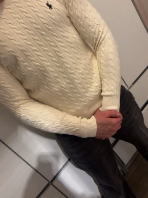 Vit kabelstickad tröja Polo Ralph Lauren - Säljer en vit kabelstickad tröja från Polo Ralph Lauren med klassisk rund hals och broderad logga på bröstet. Tröjan har långärmad passform och ribbade muddar vid ärmslut och nederkant. Perfekt för dig som gillar stilrena och tidlösa plagg.