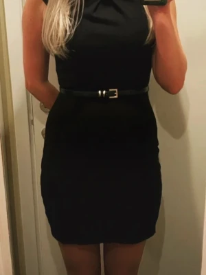 Marinblå klänning från Warehouse - Snygg marinblå kortklänning från Warehouse i storlek 36. Klänningen har en figurnära passform, korta ärmar och en dekorativ detalj vid halsen. Den stängs med dragkedja bak och har ett stilrent, enfärgat utseende. Perfekt för dig som gillar klassisk och enkel stil.            Bältet ingår inte