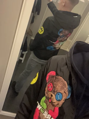 Svart vindjacka med björntryck - Säljer en svart vindjacka med färgglatt björntryck på både fram- och baksidan. Jackan har huva, dragkedja framtill och en gul smiley-detalj på ärmen. Materialet är troligtvis polyester och jackan har en normal passform. Perfekt för dig som gillar streetwear och unika prints. Jackan drar till sig extremt mycket blickar för folk som gillar o vara i centrum och ovanligt mycket komplimanger för en så färgglad jacka passar lerfejt till hösten och sommaren.