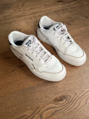 Vita Reebok sneakers med platt sula - Säljer ett par klassiska vita Reebok sneakers med svart detalj på hälen och Reebok-logga på sidan och bak. Skorna har snörning, rund tå och platt sula i syntetmaterial. Perfekt för dig som gillar stilrena och tidlösa sneakers.