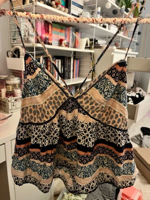 Mönstrad linnetopp med smala axelband - Säljer en färgglad linnetopp med smala axelband och flera olika mönster i blått, beige, svart och orange. Toppen har ett bohemiskt uttryck med blandade grafiska och blommiga mönster, samt en lätt v-ringad front. Perfekt för varma dagar och sommarhäng.