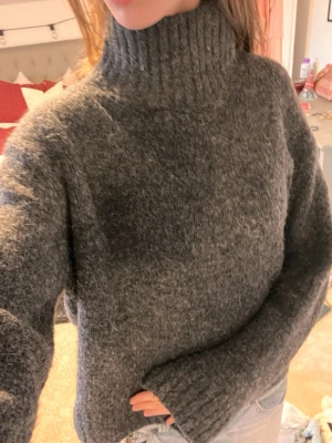 Mörkgrå stickad polotröja - Mysig mörkgrå stickad polotröja med hög krage och långa ärmar. Tröjan har en loose fit och är perfekt för kyliga dagar. Materialet känns fluffigt och värmande, och muddar finns vid både ärmslut och nederkant. Enkel att styla med jeans eller kjol.