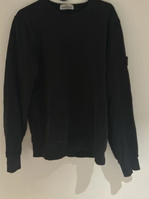 Svart sweatshirt från Stone Island - Svart sweatshirt från Stone Island med klassisk rund hals och ribbade muddar. Tröjan har lång ärm och den ikoniska Stone Island-loggan på vänster ärm. Materialet är mjuk bomull som känns skön mot huden. Perfekt för en chill och stilren look. Tröjan är använd några gånger försiktigt men är tyvärr för liten för mig. Det är storlek XL i juniorstorlek vilket passar S i herrstorlek. Hör av dig om det är något du undrar över!