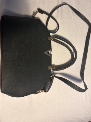 Svart axelväska med gulddetaljer - Snygg svart axelväska med guldfärgade metalldetaljer och ett V-format spänne framtill. Väskan har både handtag och en lång axelrem easily att bära crossbody. Utsidan är i strukturerat material och insidan har flera fack samt dragkedja för smart förvaring.