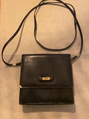 Svart axelväska från Baron i skinn - Snygg svart axelväska från Baron i äkta skinn. Väskan har en stilren, fyrkantig form med guldfärgade detaljer och ett smalt axelband. Perfekt för dig som gillar klassisk och minimalistisk design. Flera fack inuti för bra organisering.