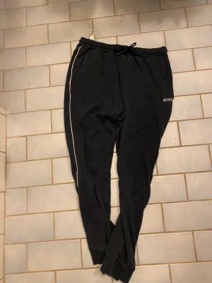 Svarta mjukisbyxor från BOSS - Svarta mjukisbyxor från BOSS med vit piping längs sidorna och logga broderad på vänster ficka. Byxorna har resår och snörning i midjan samt muddar vid bensluten. Perfekta för chill eller träning.