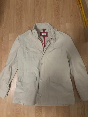 Beige field jacka från Brunello Cucinelli - Snygg beige field jacka från Brunello Cucinelli med röda detaljer på insidan i storlek 50 passar en L/M. Jackan har fyra stora fickor framtill, dragkedja och tryckknappar. Tillverkad i Italien och har justerbar midja med snörning. Perfekt för dig som gillar stilrena och exklusiva plagg.