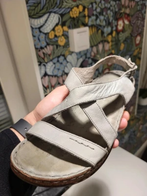 Copenhagen dream comfort skinnsandaler med korksula 38 - Säljer ett par ljusgrå sandaler i mjukt skinn med breda remmar och justerbart spänne vid hälen. Skorna har en rejäl platåsula i kork-look som ger extra höjd och en avslappnad vibe. Perfekta för sommardagar och passar till både jeans och klänning.