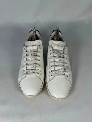 Vita Saint Laurent sneakers i skinn - Säljer ett par klassiska vita sneakers från Saint Laurent Paris. Skorna är tillverkade i slätt skinn med rund tå, vita snören och diskret logga på plösen. Snygg minimalistisk design med låg profil och neutrala detaljer. Perfekta till jeans eller kjol. Kommer med box och påse till. 