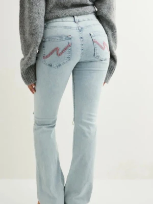 Ljusblå bootcut jeans med slitningar - Säljer ett par ljusblå jeans med bootcut passform och slitna detaljer över knäna. Jeansen har klassisk femficksdesign och coola broderade detaljer i rött på bakfickorna. Perfekta för en avslappnad och trendig look. Jag har bara testat jeansen på en gång.