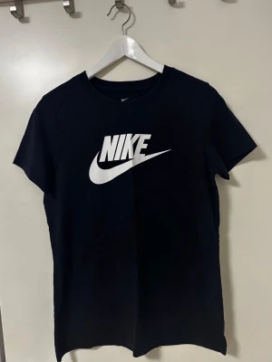 Svart Nike t-shirt med logga - Svart t-shirt från Nike med stor vit logga framtill. Klassisk rund halsringning och korta ärmar. Tillverkad i mjuk bomull som är skön att ha på sig. Perfekt för dig som gillar sportiga och stilrena plagg.